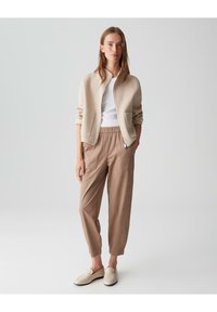 Beiger Bomberjacke mit zwei Taschen, weißem T-Shirt, taubfarbenen tapered Hosen und cremefarbenen Loafers. Glatte Textur, lockere Passform und neutrale Farben.