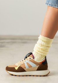 Zapatilla colorida con un upper en tonos beige y marrón, acentos azules y una suela blanca. Llevada con un calcetín amarillo texturizado.