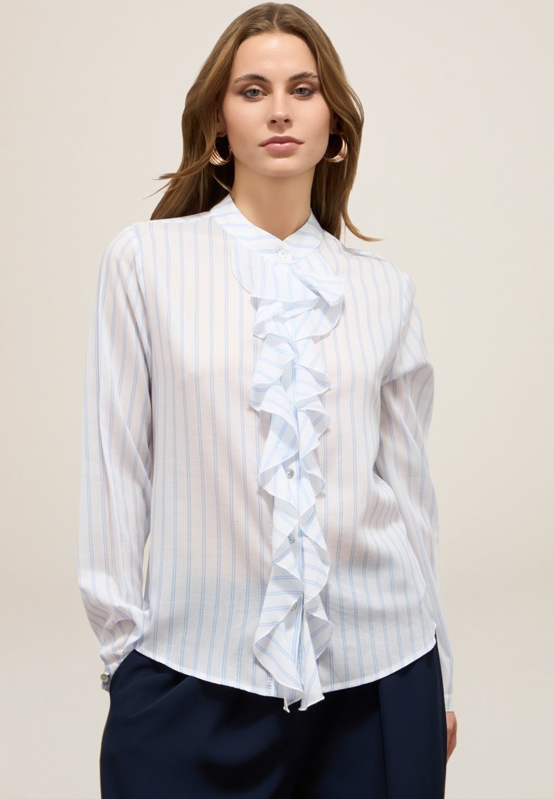 Blusa bianca a maniche lunghe con strisce verticali blu e un dettaglio centrale di rouches con bottoni, realizzata in tessuto leggero.