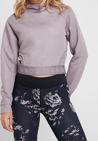 Pitkähihaisen vaalean violetin crop-topin, jossa on korkea kaulus, tekstuuriset sivut ja "UNDER ARMOUR" -logo helmassa; yhdistetty mustiin kukkaleggingseihin.