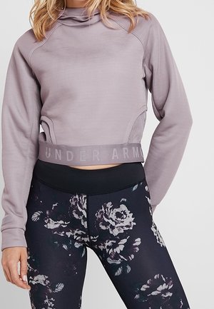 Langarm crop top in lichtpaars met hoge kraag, geprofileerde zijkanten en het "UNDER ARMOUR" logo aan de onderkant; gecombineerd met zwarte bloemenlegging.