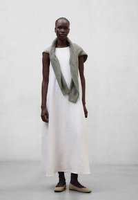 Femme portant une robe maxi blanche sans manches avec un pull en maille vert drapé sur les épaules et des ballerines beiges avec des chaussettes noires.