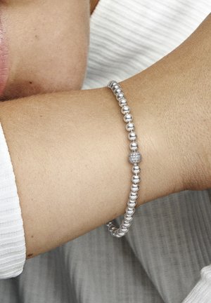 Silberperlenarmband mit glatten, sphärischen Perlen und einem strukturierten, dekorativen Element. Am Handgelenk getragen, vor einem hellen Hintergrund.