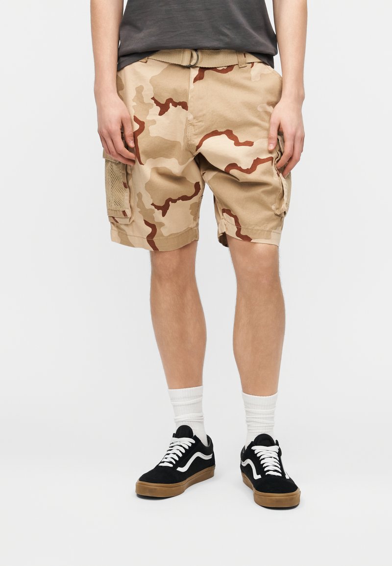 Άτομο που φοράει μπεζ και καφέ καμουφλαρισμένα σορτς με τσέπες, άσπρες κάλτσες και μαύρα αθλητικά παπούτσια με λευκά κορδόνια και σόλες, στέκεται σε απλό φόντο.