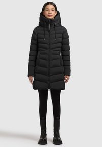 Črn puffer plašč s kapuco, z zadrgo spredaj in dodatki na vrvico. Ima prešito teksturo in dolžino do kolen. Nosljiv s črnimi leginami in čevlji.