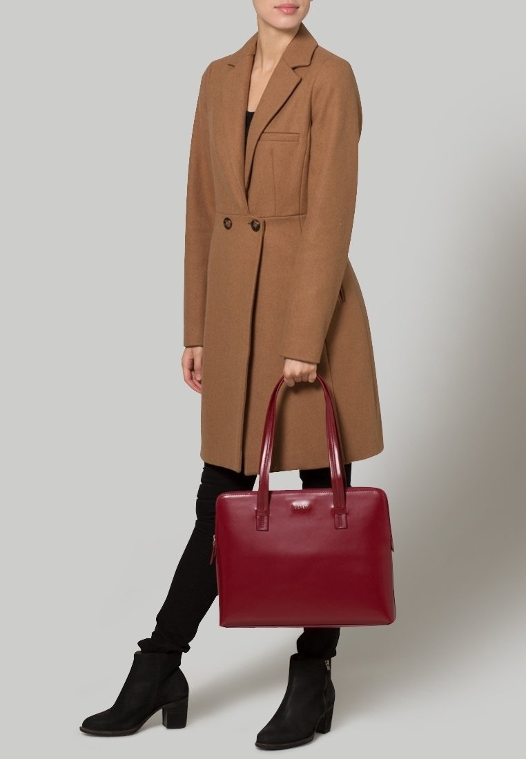 Femme portant un manteau marron, un pantalon noir et des bottines noires tenant un grand sac cabas en cuir rouge sur un fond gris.
