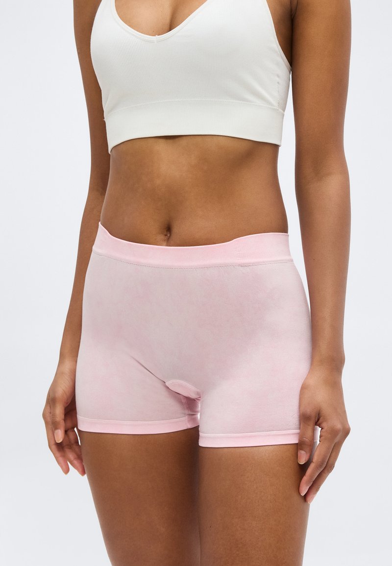 Femme portant un soutien-gorge de sport blanc et un boxer sans couture rose clair, debout devant un fond uni.
