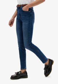 Jeans ajustados de color azul con una cintura alta, que presentan un diseño clásico de cinco bolsillos y un ligero desgastado, combinados con zapatos negros y un cinturón trenzado.