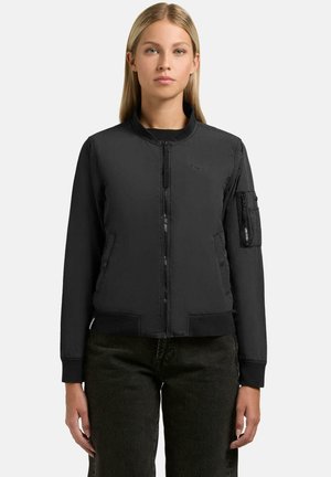 Blondynka ubrana w czarną bomberkę na suwak z prążkowanym kołnierzem, mankietami i krawędzią oraz z zasuwaną kieszenią na lewej rękawie, w połączeniu z ciemnymi spodniami.