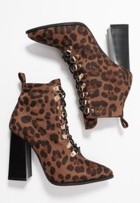 Bottes chevilles à talons hauts avec imprimé léopard marron, lacets noirs, œillets dorés, bouts pointus et talons épais noirs sur fond blanc.