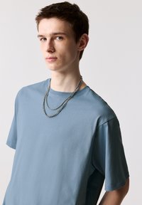 Lyseblå bomulds t-shirt med rund halsudskæring og korte ærmer, suppleret med lagte sølvkædehalskæder. Flad tekstur, minimalistisk design.