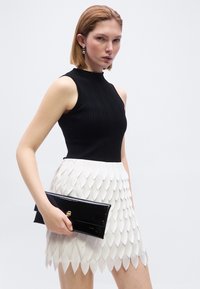 Femme portant un haut noir sans manches et une jupe blanche à texture pétale, tenant une pochette noire brillante avec un fermoir doré.