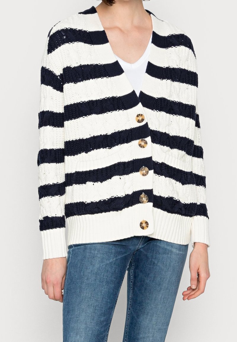 Personne portant un cardigan tricoté à rayures horizontales blanches et noires avec cinq boutons, par-dessus une chemise blanche et un jean bleu.