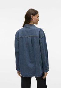 Dark blue denim oversized shirt with a back pleat, a pointed collar, and button cuffs. Smooth texture with visible stitching details.