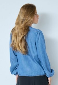 Blusa in denim azzurro chiaro con maniche lunghe, spalle arricciate e una vestibilità leggermente ampia. Presenta un collo rotondo e un orlo dritto.
