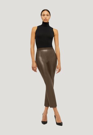 Femme debout devant un fond uni, portant un col roulé sans manches noir, un pantalon en cuir marron et des talons hauts noirs.