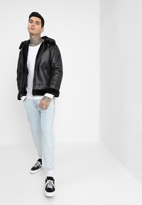 Chaqueta bomber de cuero negra con forro de piel sintética, que presenta un cierre de cremallera y botón. Combinada con vaqueros azul claros y zapatillas negras.