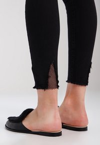 Vaqueros de mezclilla negra con dobladillo deshilachado, con recortes que revelan tela de malla. Combinados con sandalias planas negras con un detalle texturizado en la tira.