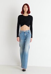 Crni dugi rukav crop top s uvijenim detaljem; svijetloplave široke traperice s izblijedjelim uzorkom; model nosi otvorene crne cipele.