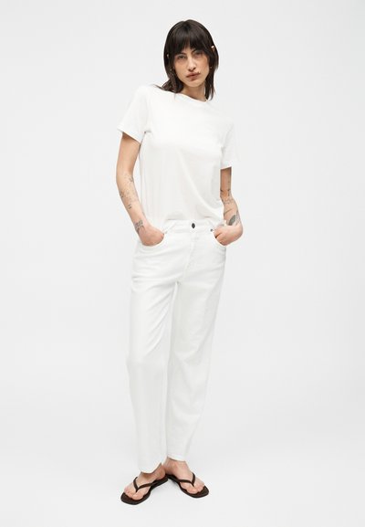 Γυναίκα με σκούρα μαλλιά που φοράει απλό λευκό μπλουζάκι, λευκό παντελόνι και μαύρα σαγιονάρες, στέκεται με τα χέρια στις τσέπες σε λευκό φόντο.