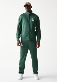 Πράσινη φόρμα από απαλό ύφασμα, με ζακέτα με φερμουάρ που έχει πλευρικές ρίγες και λογότυπο, συνδυασμένη με παντελόνι στο ίδιο ύφος και άσπρα αθλητικά παπούτσια.