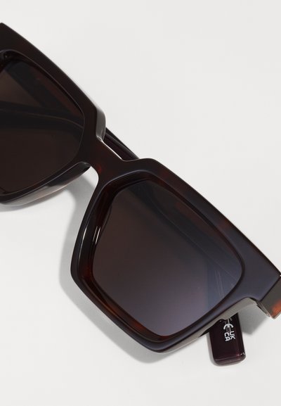 Dark brown oversized sunglasses with a thick plastic frame, featuring dark lenses and a glossy finish. Accented with a subtle logo on the arm.