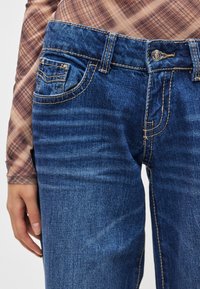 Μπλε τζιν με ελαφρύ βουρτσισμό, κίτρινη ραφή, εμπρόσθια τσέπη και κούμπωμα με κουμπί· φορεμένο με μακρυμάνικο πουκάμισο καρό.