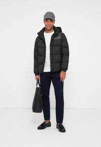 Sort puff jakke med hætte, quilted design, båret over en hvid skjorte. Marineblå bukser, sorte sko og en grå flad cap fuldender outfittet.