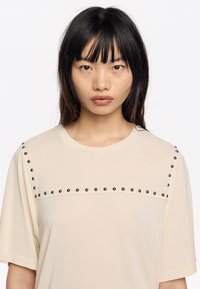Camiseta beige de manga corta con un panel texturizado, adornada con tachuelas negras a lo largo de la costura del hombro y la zona del pecho. Tejido suave.