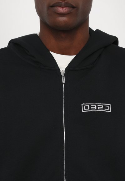 Black zip-up hoodie with a soft texture, featuring a drawstring hood and a white "CESO" logo on the left chest. Silver zipper detail visible.