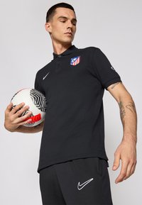 Sort polo-shirt med et Nike-logo og Atletico Madrid-crest, parret med sorte bukser med et hvidt Nike-swoosh, mens han holder en fodbold.