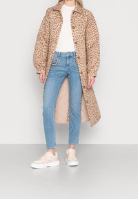 Béžový dlouhý kabát s leopardím potiskem a zapínáním na knoflíky, kombinovaný se světle modrými džínami a teniskami na platformě. Hladká látka, volný střih.