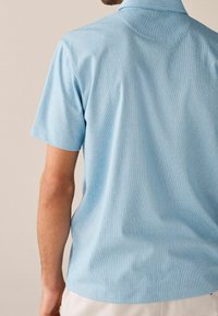 Camisa de manga corta con cuello en azul claro, con tejido texturizado y rayas sutiles, vista trasera, superficie lisa y ajuste relajado.