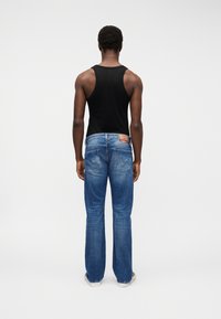 Άνδρας που φοράει μαύρο αμάνικο μπλουζάκι, μπλε τζιν με δερμάτινη ετικέτα στη μέση και λευκά παπούτσια, στέκεται πλάτη σε λευκό φόντο.