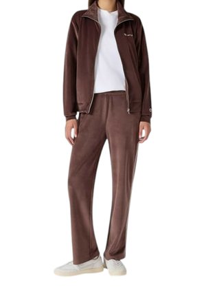 Personne portant une veste zippée en velours marron et un pantalon assorti avec un t-shirt blanc et des baskets blanches, debout face à l'avant.