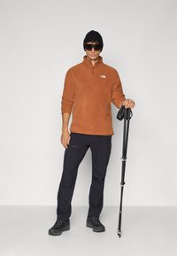 Pull en polaire marron avec un col montant et une fermeture éclair quart, pantalon noir, bottes de randonnée noires et un bonnet noir, tenant un bâton de randonnée.