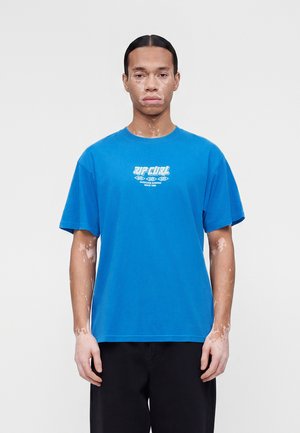 Fiatal személy vitiligóval, kék Rip Curl pólót és fekete nadrágot visel, egyszínű világos háttér előtt áll.