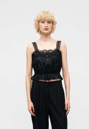 Personne blonde portant un haut noir sans manches à boutons, orné de dentelle, et un pantalon taille haute noir, debout devant un fond uni.