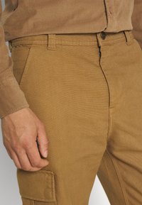 Rudi cargo kelnaičiai su tekstūruotu audiniu, turintys priekinį užtrauktuką, diržo kilpas ir šoninį kišenę. Artimas vaizdas, kuriame ranka remiasi į klubus.