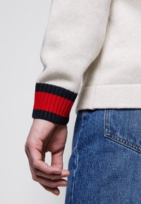 Mano rilassata a fianco, indossando un maglione a maglia crema con strisce nere e rosse sui polsini, abbinato a jeans in denim blu.