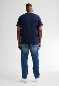 Námořnická modrá bavlněná trička s krátkým rukávem, spárovaná se světlými modrými denimovými džíny. Úzký střih, casual styl, bílé tenisky viditelné na spodní části.