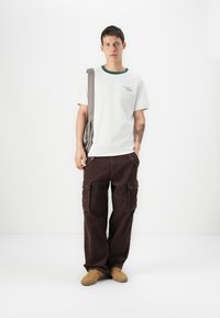 Camiseta blanca de manga corta con cuello verde, pantalones cargo marrones con bolsillos y zapatillas beige. El modelo lleva un bolso de hombro gris.