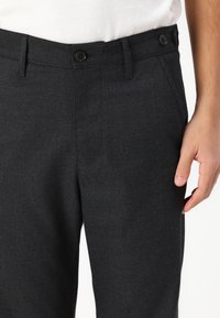 Pantalon gris foncé en tissu texturé, présentant un devant plat, deux boutons et des poches latérales. Main placée sur la taille.