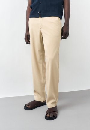 Dolná časť tela osoby, ktorá nosí béžové nohavice rovného strihu, tmavomodrú košeľu na gombíky a tmavohnedé sandále s krížovými remienkami, stojaca na šedej podlahe.