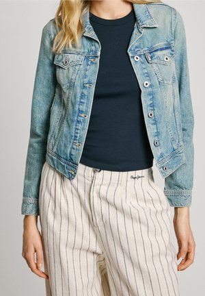 Femme portant une veste en jean bleu clair par-dessus une chemise noire et un pantalon blanc à fines rayures verticales noires, les mains détendues le long du corps.