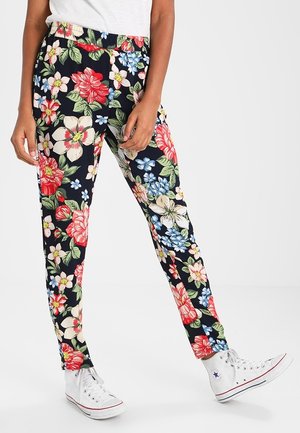 Osoba ubrana w granatowe spodnie w kwiaty w kolorach czerwonym, białym i niebieskim, zestawione z białymi wysokimi trampkami i białą bluzką.