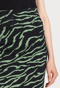 Falda lápiz negra con un patrón de rayas de tigre verde. Tejido suave con una silueta ajustada. Diseño de cinturilla mostrado en la parte superior.