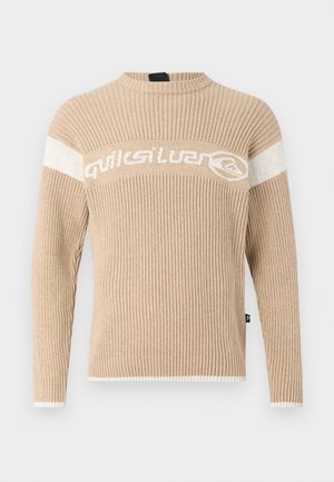 Pull en beige à côtes avec un col haut, présentant des rayures blanches contrastées et un logo sur la poitrine. Texture douce, manches longues.