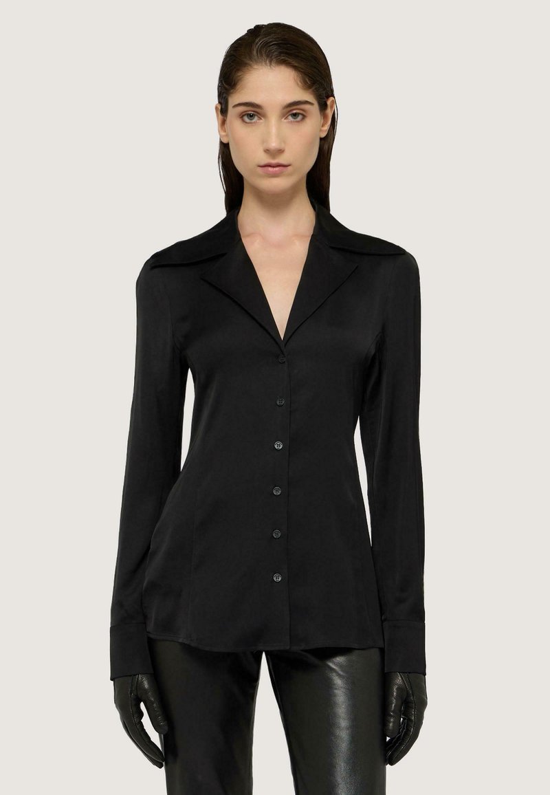 Blusa nera a maniche lunghe con colletto a punte, chiusura frontale con bottoni, texture liscia e silhouette aderente, abbinata a pantaloni neri lucidi.