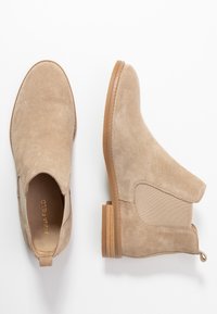 Bottines Chelsea en daim beige avec panneaux latéraux élastiques, languette de tirage au talon et petit talon en bois empilé, vues de dessus et de côté.